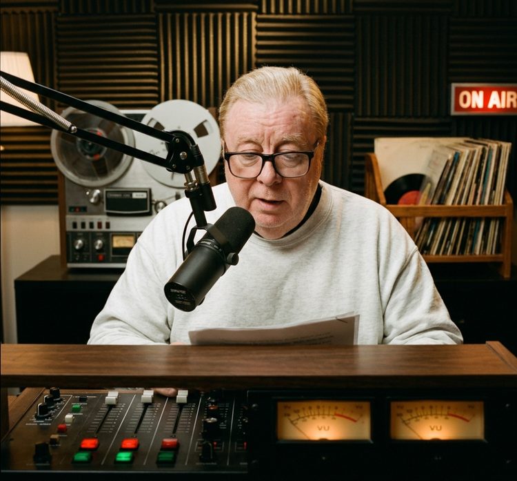 Thomas Coss at the broadcast console in his studio with ON AIR sign, reel-to-reel tape deck, vinyl records, and VU meters